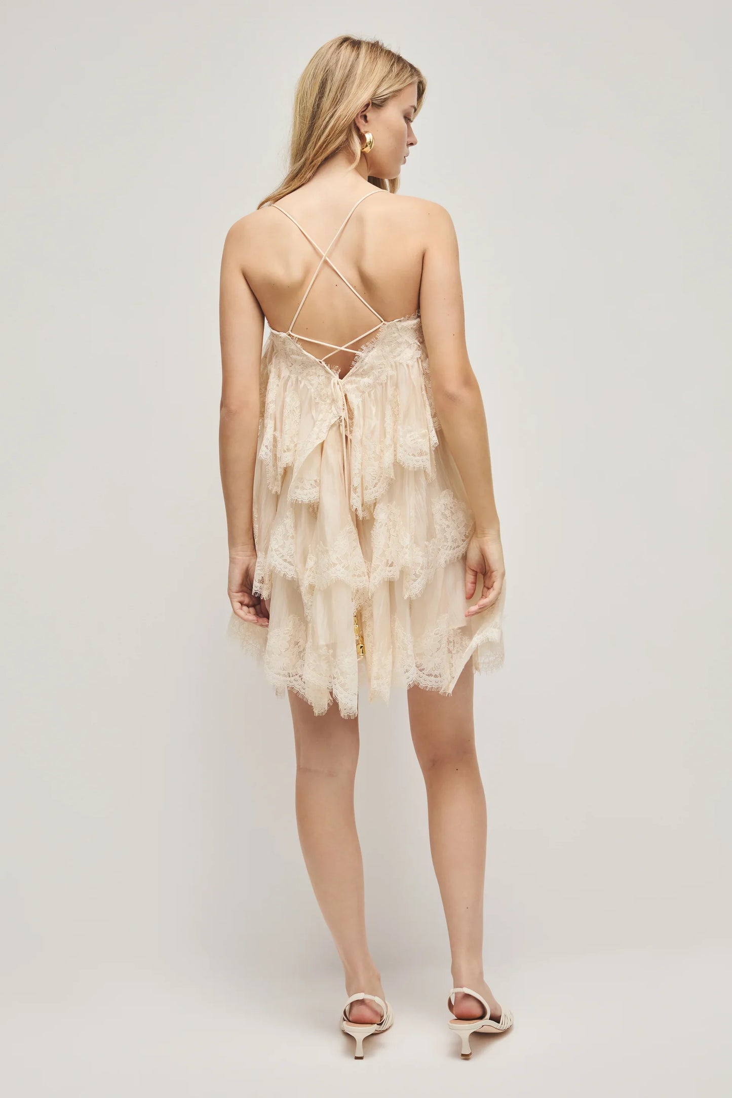 A woman models a cream-coloured, tiered mini dress featuring semi-sheer ruffles, delicate lace trim, and thin spaghetti straps.