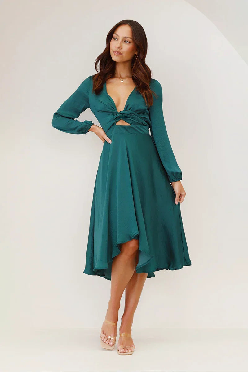 As shown - A woman models a long-sleeved, emerald green satin midi dress against a white background. The dress has a deep V-neck with a large tie-front bow, a matching tie-belt at the waist, and an A-line skirt.