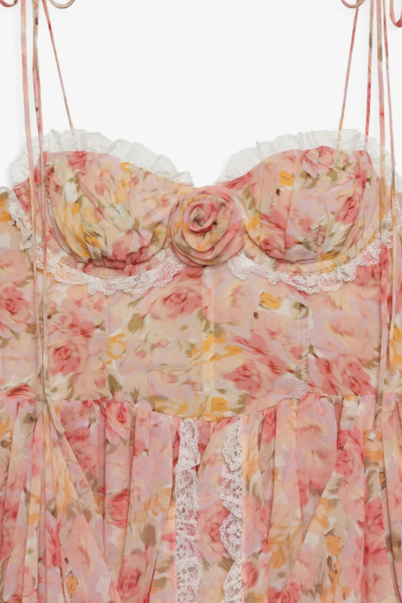 Elea - A woman models a romantic floral mini dress featuring a pale pink and yellow rose print. The dress has a structured corset bodice with a sweetheart neckline, delicate tie-shoulder straps, a rosette appliqué at the bust, and a layered, ruffled asymmetrical skirt trimmed with white lace.