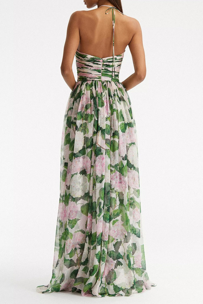 A woman modelling a flowing maxi dress with a ruched halterneck bodice and a large pink and green botanical floral print.