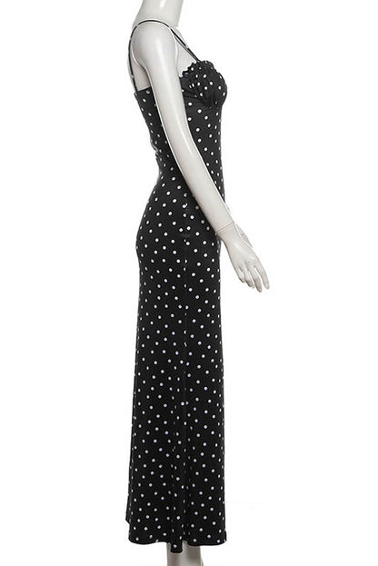 Black - A woman standing by the water wearing a black maxi dress with white polka dots, featuring spaghetti straps and a ruched bodice.