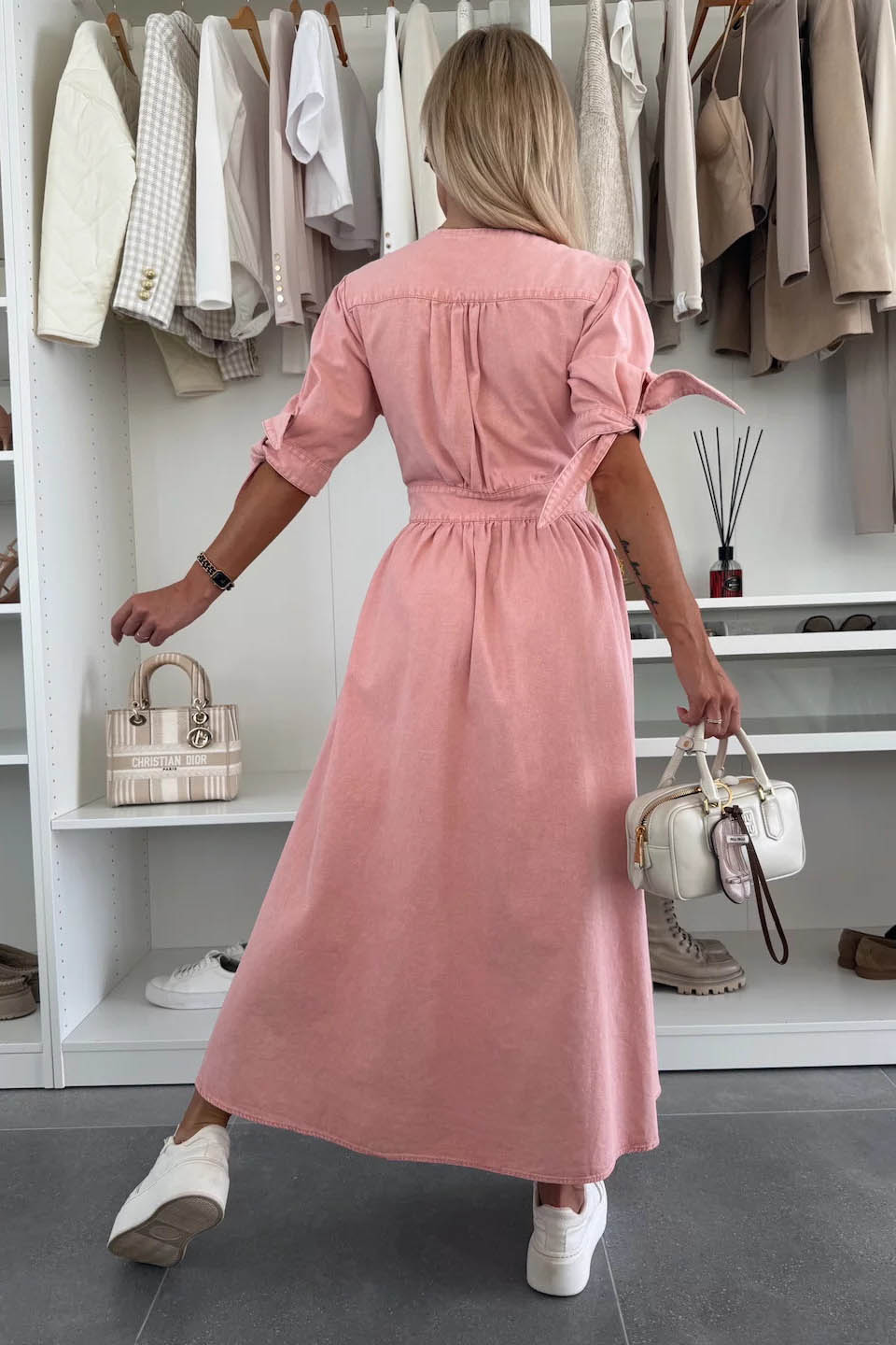 Pink - Woman wearing a light blue denim-look midi dress with a V-neck, button-down front, and tie-detail short sleeves, styled with trainers and a small white handbag.
