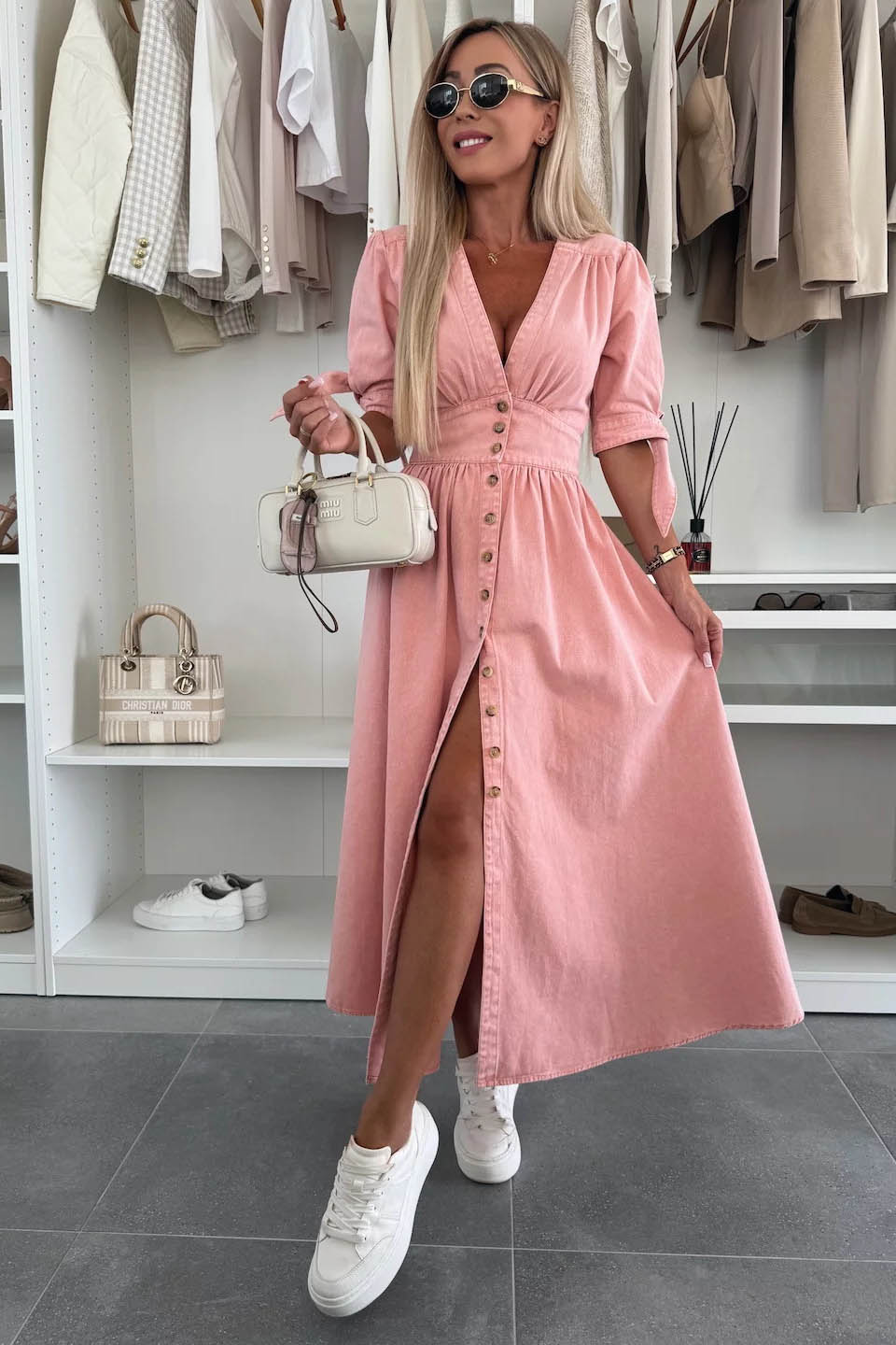 Pink - Woman wearing a light blue denim-look midi dress with a V-neck, button-down front, and tie-detail short sleeves, styled with trainers and a small white handbag.