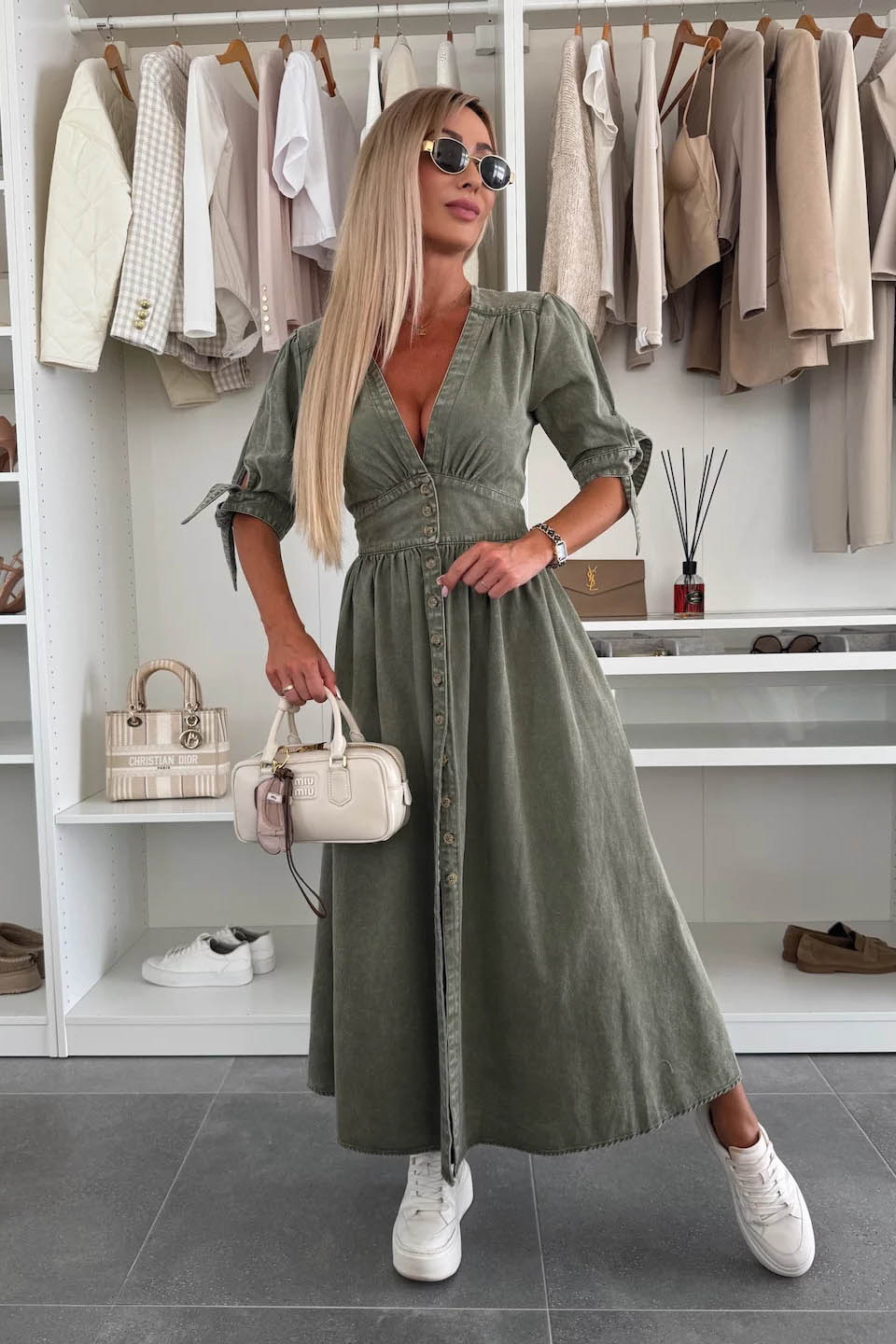 Green - Woman wearing a light blue denim-look midi dress with a V-neck, button-down front, and tie-detail short sleeves, styled with trainers and a small white handbag.