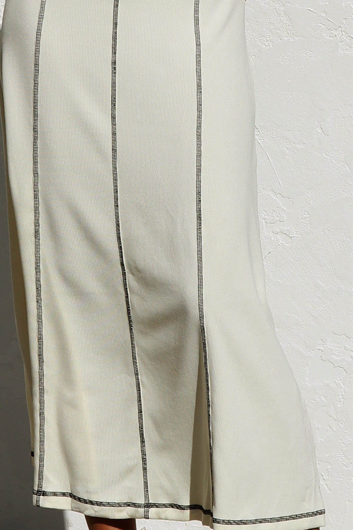 White - A woman stands against a white wall wearing a sleeveless, form-fitting, off-white midi dress. The dress has a high neckline and is detailed with dark vertical contrast stitching along the seams and armholes.