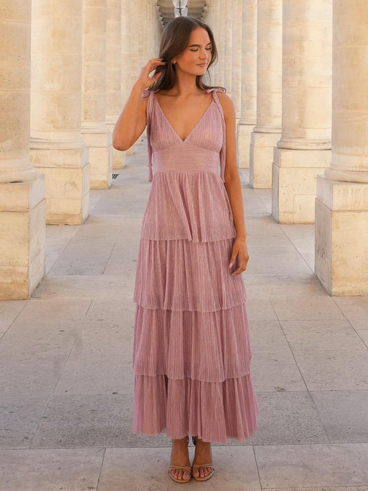A woman stands in a stone colonnade wearing a dusty pink, shimmering maxi dress featuring a V-neck, tie-shoulders, a ruched waist, and a long, tiered skirt.