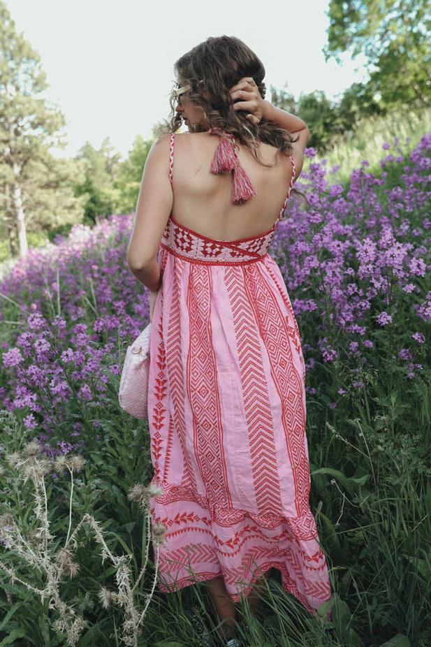 Woman standing in a field of purple flowers wearing a pink and red halter maxi dress with a geometric print, embroidered bodice, and tiered skirt.