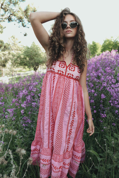 Woman standing in a field of purple flowers wearing a pink and red halter maxi dress with a geometric print, embroidered bodice, and tiered skirt.