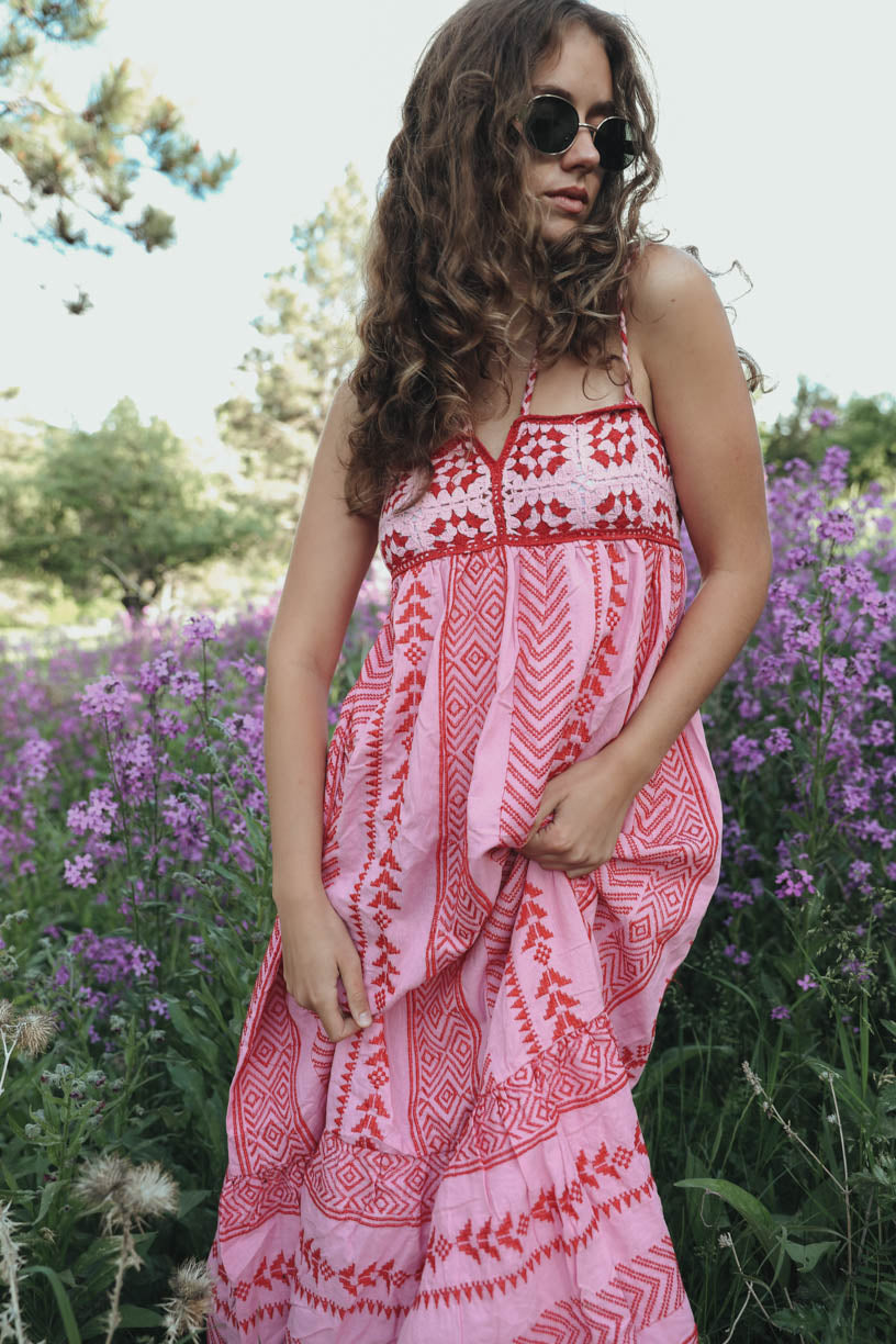 Woman standing in a field of purple flowers wearing a pink and red halter maxi dress with a geometric print, embroidered bodice, and tiered skirt.