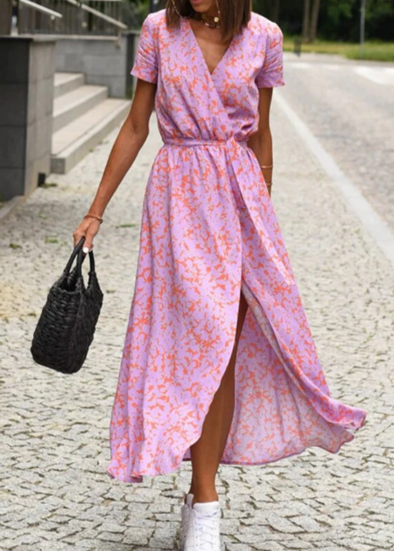 Pink - A woman walking outdoors wearing a vibrant pink and green floral print midi dress with a V-neckline, short sleeves, a tie waist, and a front slit.