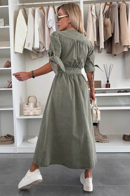 Green - Woman wearing a light blue denim-look midi dress with a V-neck, button-down front, and tie-detail short sleeves, styled with trainers and a small white handbag.