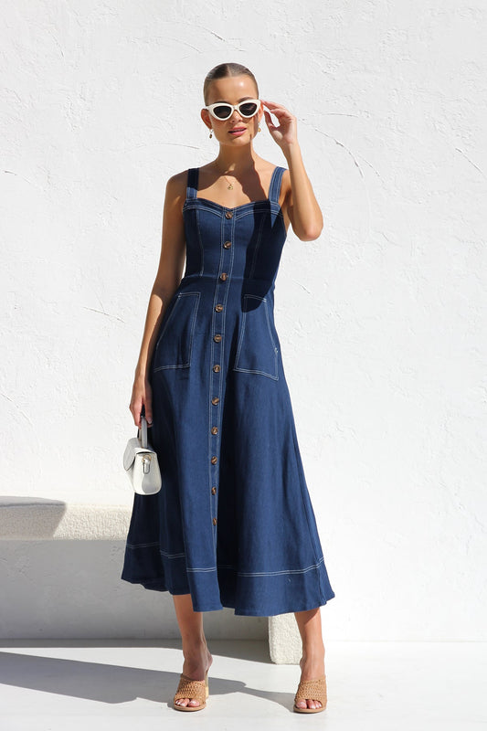 A woman models a dark blue, sleeveless A-line midi dress with contrast white topstitching and a full button-front. She wears white cat-eye sunglasses and holds a small white handbag, standing against a white textured wall.