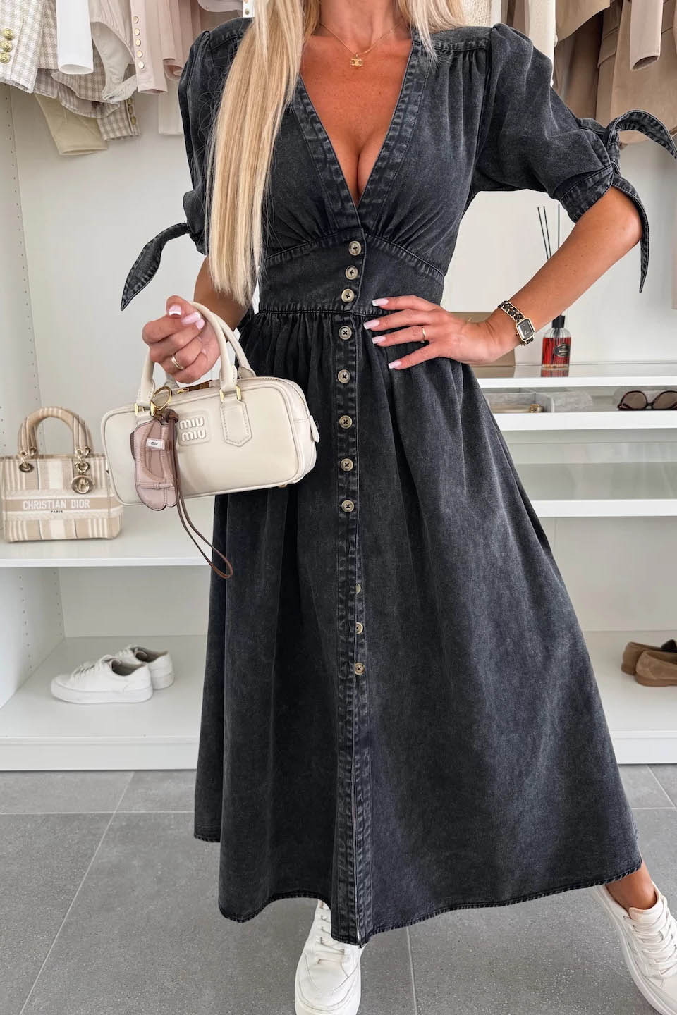 Black - Woman wearing a light blue denim-look midi dress with a V-neck, button-down front, and tie-detail short sleeves, styled with trainers and a small white handbag.