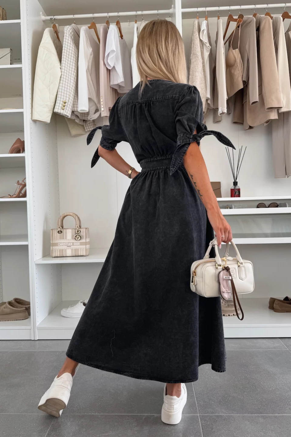 Black - Woman wearing a light blue denim-look midi dress with a V-neck, button-down front, and tie-detail short sleeves, styled with trainers and a small white handbag.