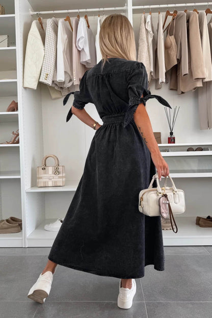 Black - Woman wearing a light blue denim-look midi dress with a V-neck, button-down front, and tie-detail short sleeves, styled with trainers and a small white handbag.