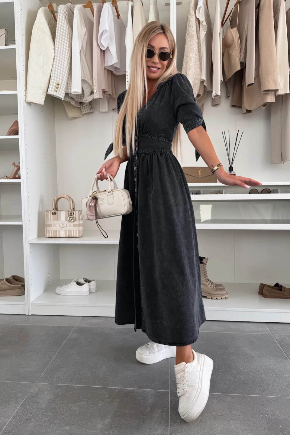 Black - Woman wearing a light blue denim-look midi dress with a V-neck, button-down front, and tie-detail short sleeves, styled with trainers and a small white handbag.