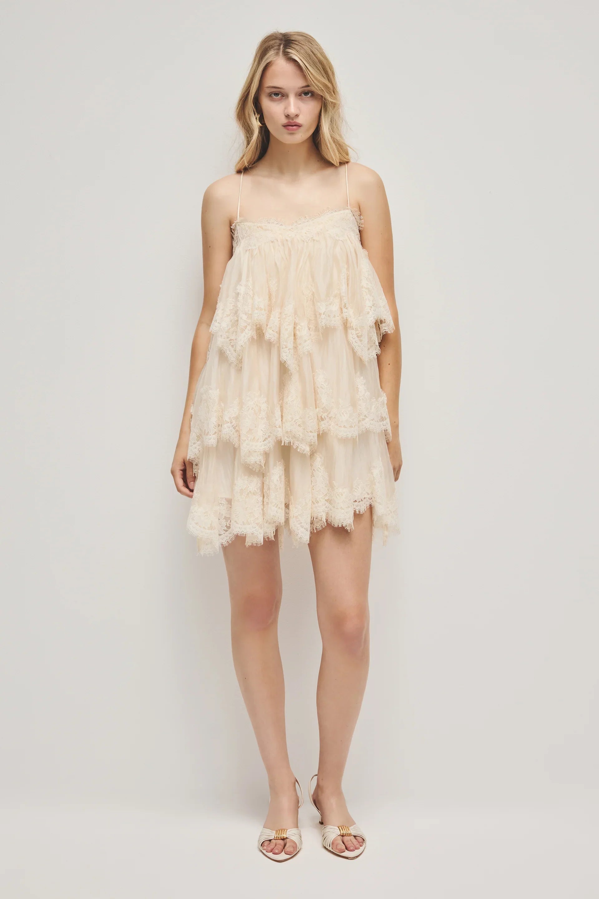 A woman models a cream-coloured, tiered mini dress featuring semi-sheer ruffles, delicate lace trim, and thin spaghetti straps.