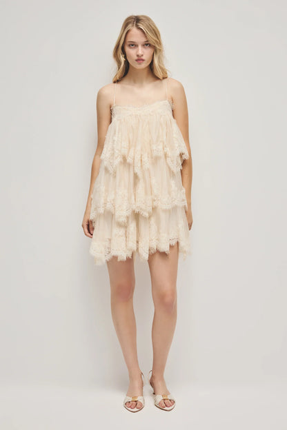 A woman models a cream-coloured, tiered mini dress featuring semi-sheer ruffles, delicate lace trim, and thin spaghetti straps.