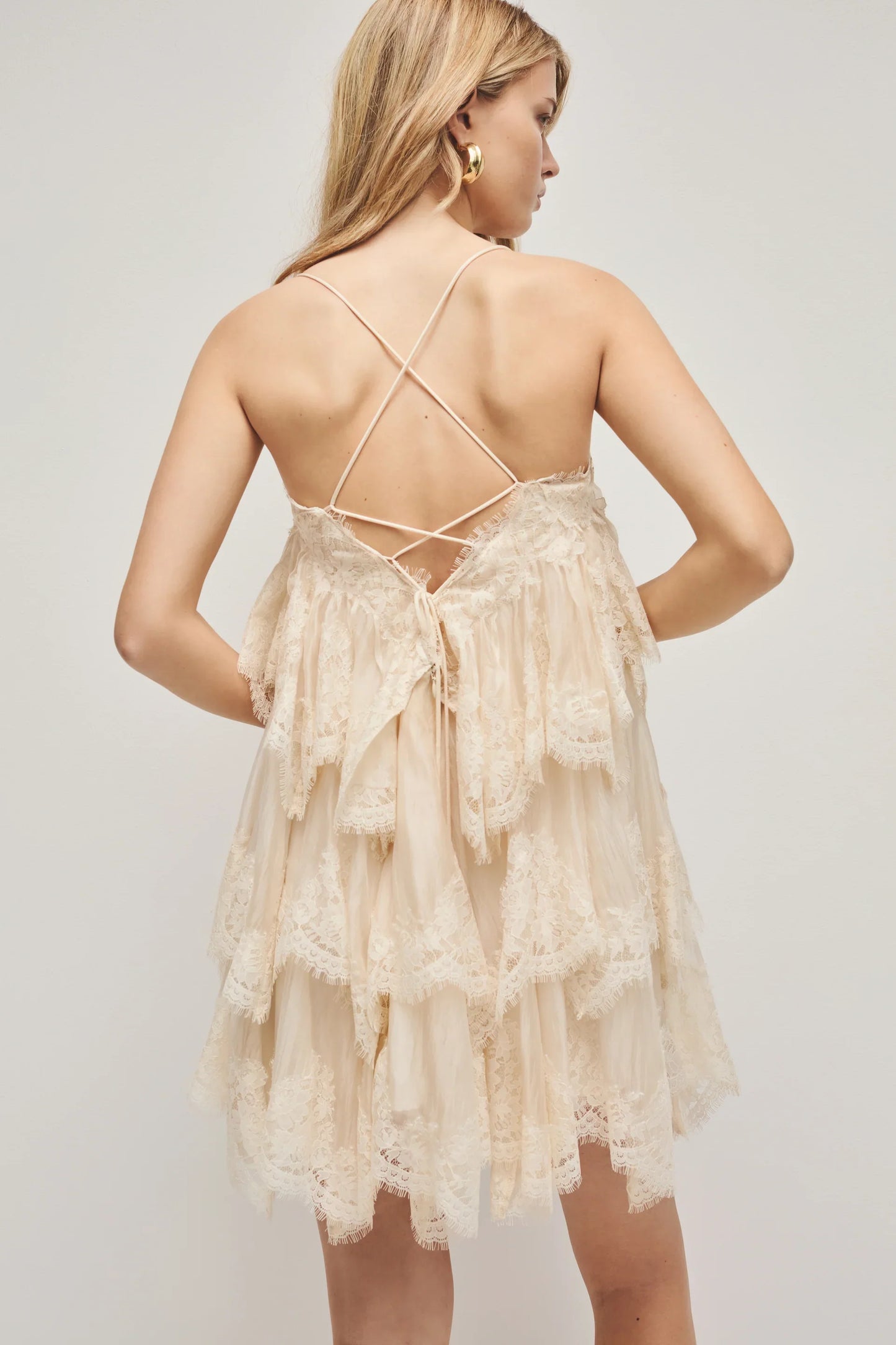 A woman models a cream-coloured, tiered mini dress featuring semi-sheer ruffles, delicate lace trim, and thin spaghetti straps.