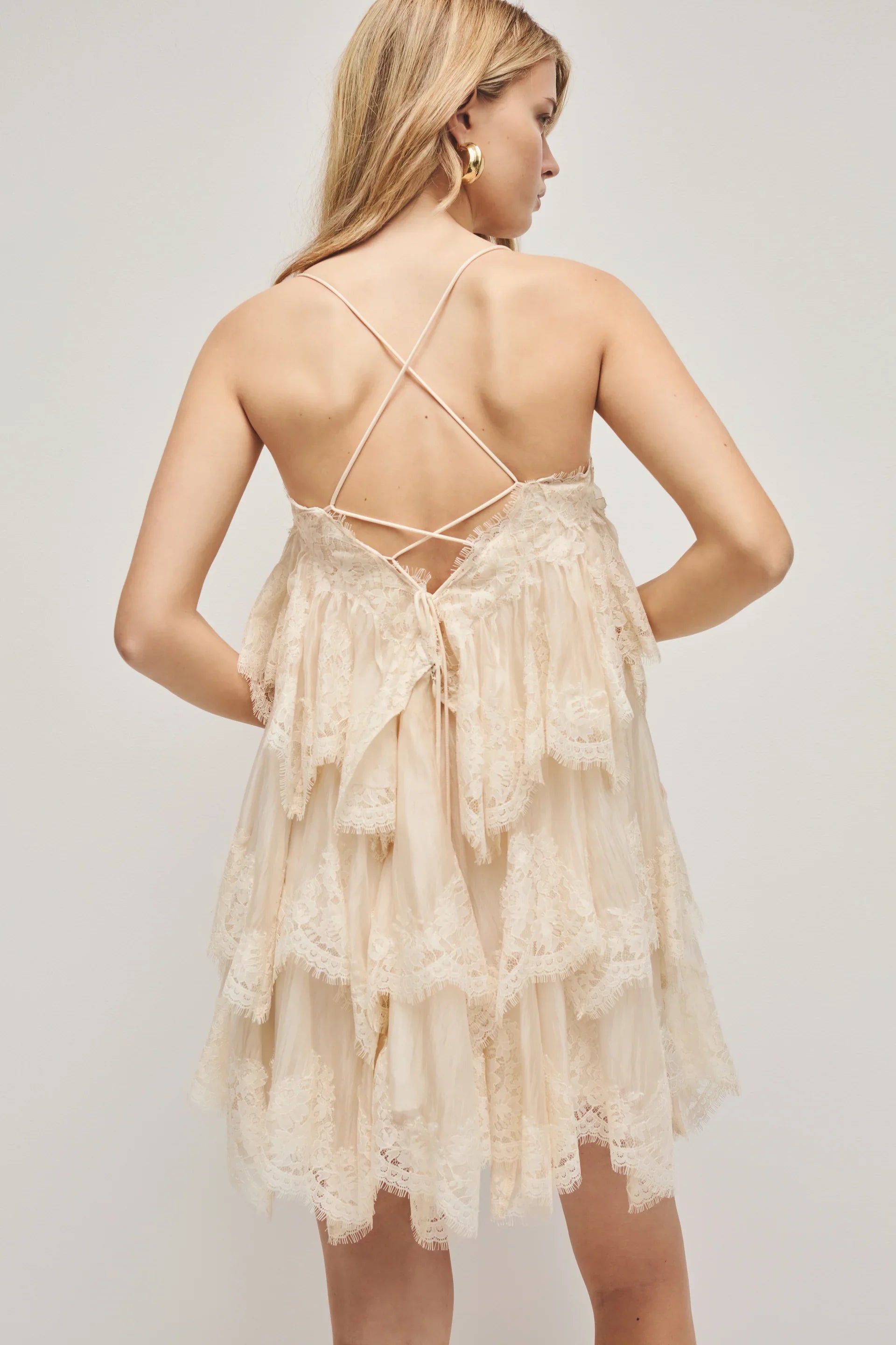 A woman models a cream-coloured, tiered mini dress featuring semi-sheer ruffles, delicate lace trim, and thin spaghetti straps.