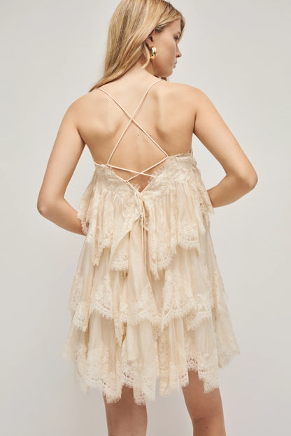 A woman models a cream-coloured, tiered mini dress featuring semi-sheer ruffles, delicate lace trim, and thin spaghetti straps.