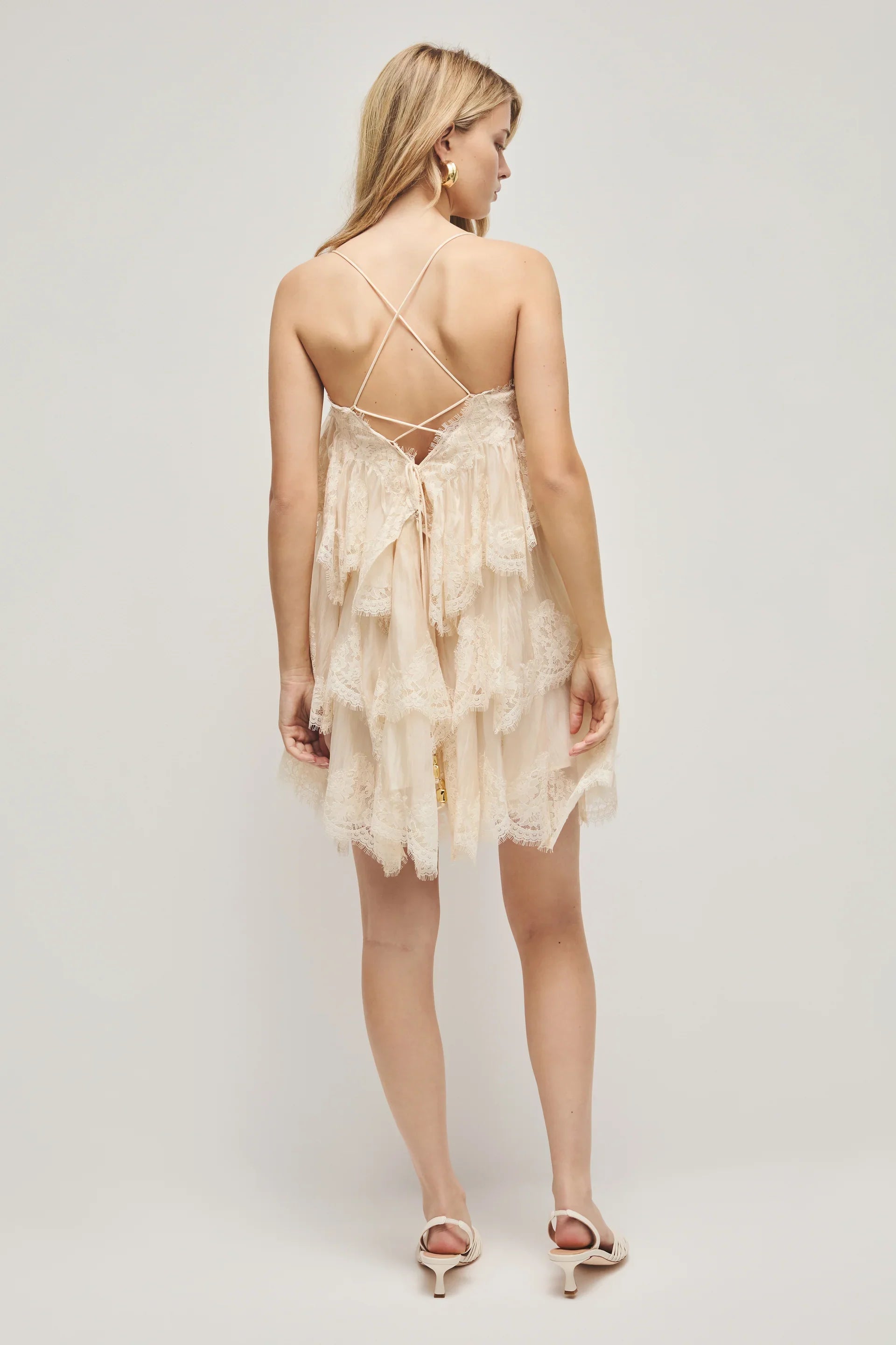 A woman models a cream-coloured, tiered mini dress featuring semi-sheer ruffles, delicate lace trim, and thin spaghetti straps.