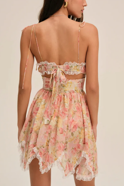 Elea - A woman models a romantic floral mini dress featuring a pale pink and yellow rose print. The dress has a structured corset bodice with a sweetheart neckline, delicate tie-shoulder straps, a rosette appliqué at the bust, and a layered, ruffled asymmetrical skirt trimmed with white lace.