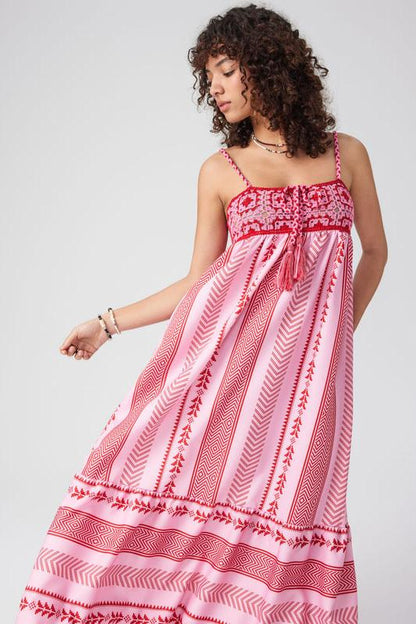 Woman standing in a field of purple flowers wearing a pink and red halter maxi dress with a geometric print, embroidered bodice, and tiered skirt.