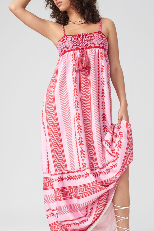 Woman standing in a field of purple flowers wearing a pink and red halter maxi dress with a geometric print, embroidered bodice, and tiered skirt.