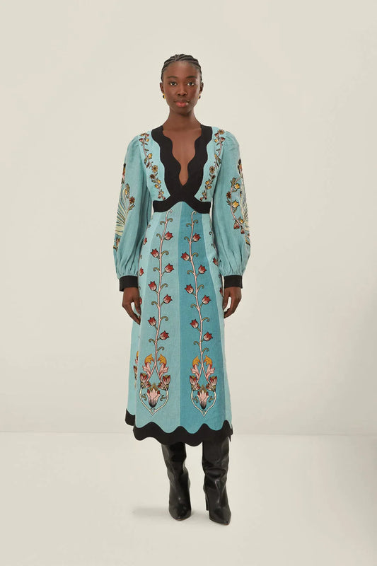 A woman models a long-sleeve, midi-length dress in shades of blue with a stylized floral print. The dress has a deep V-neckline, voluminous bishop sleeves, and a contrasting black scalloped trim at the neck, cuffs, and hem.