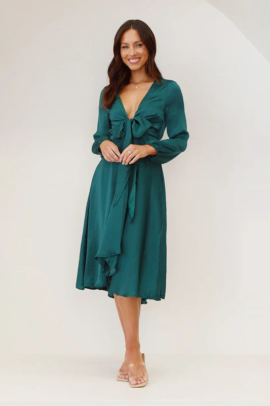 A woman models a long-sleeved, emerald green satin midi dress against a white background. The dress has a deep V-neck with a large tie-front bow, a matching tie-belt at the waist, and an A-line skirt.