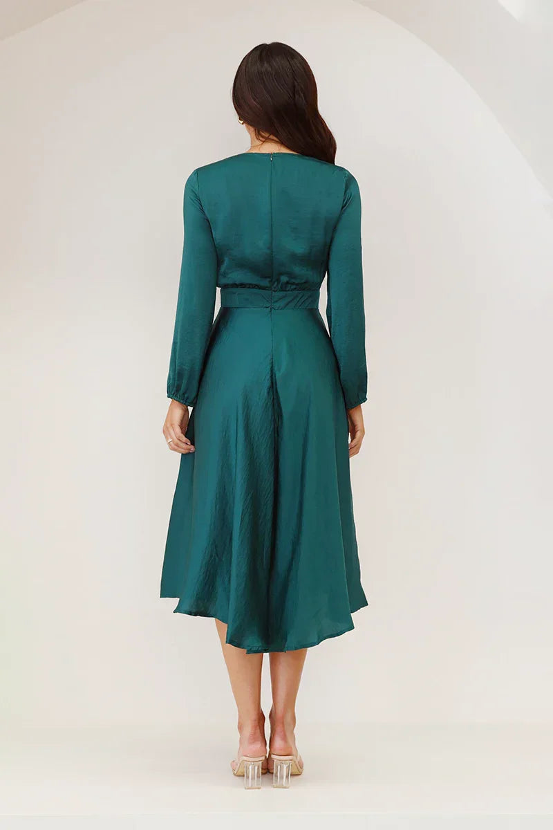 A woman models a long-sleeved, emerald green satin midi dress against a white background. The dress has a deep V-neck with a large tie-front bow, a matching tie-belt at the waist, and an A-line skirt.