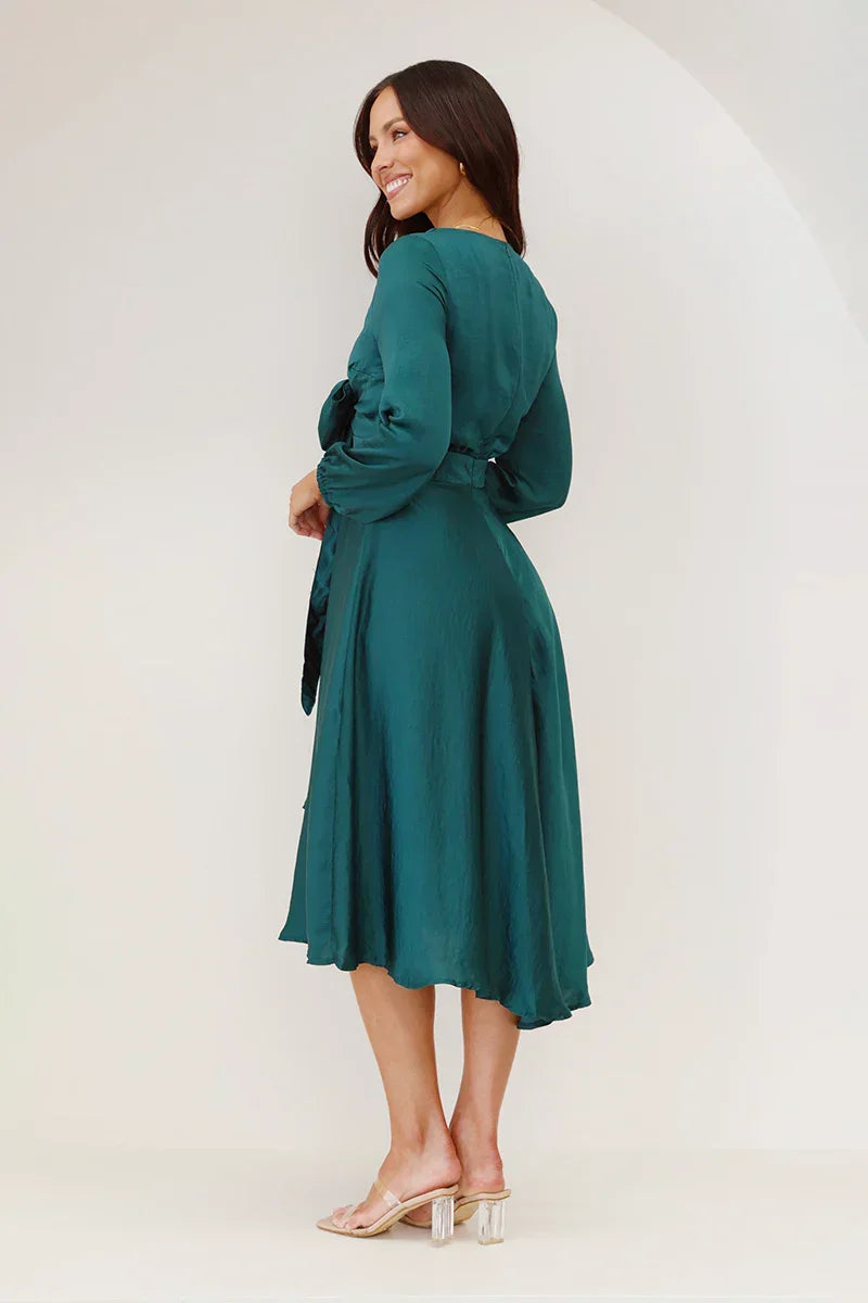 A woman models a long-sleeved, emerald green satin midi dress against a white background. The dress has a deep V-neck with a large tie-front bow, a matching tie-belt at the waist, and an A-line skirt.