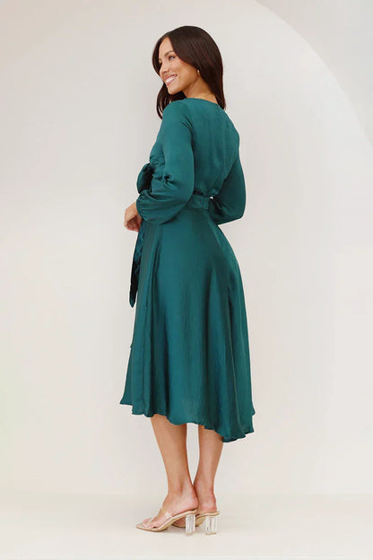 A woman models a long-sleeved, emerald green satin midi dress against a white background. The dress has a deep V-neck with a large tie-front bow, a matching tie-belt at the waist, and an A-line skirt.