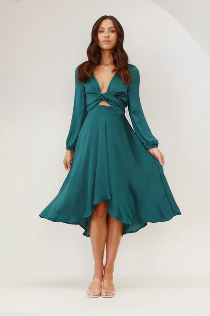 As shown - A woman models a long-sleeved, emerald green satin midi dress against a white background. The dress has a deep V-neck with a large tie-front bow, a matching tie-belt at the waist, and an A-line skirt.