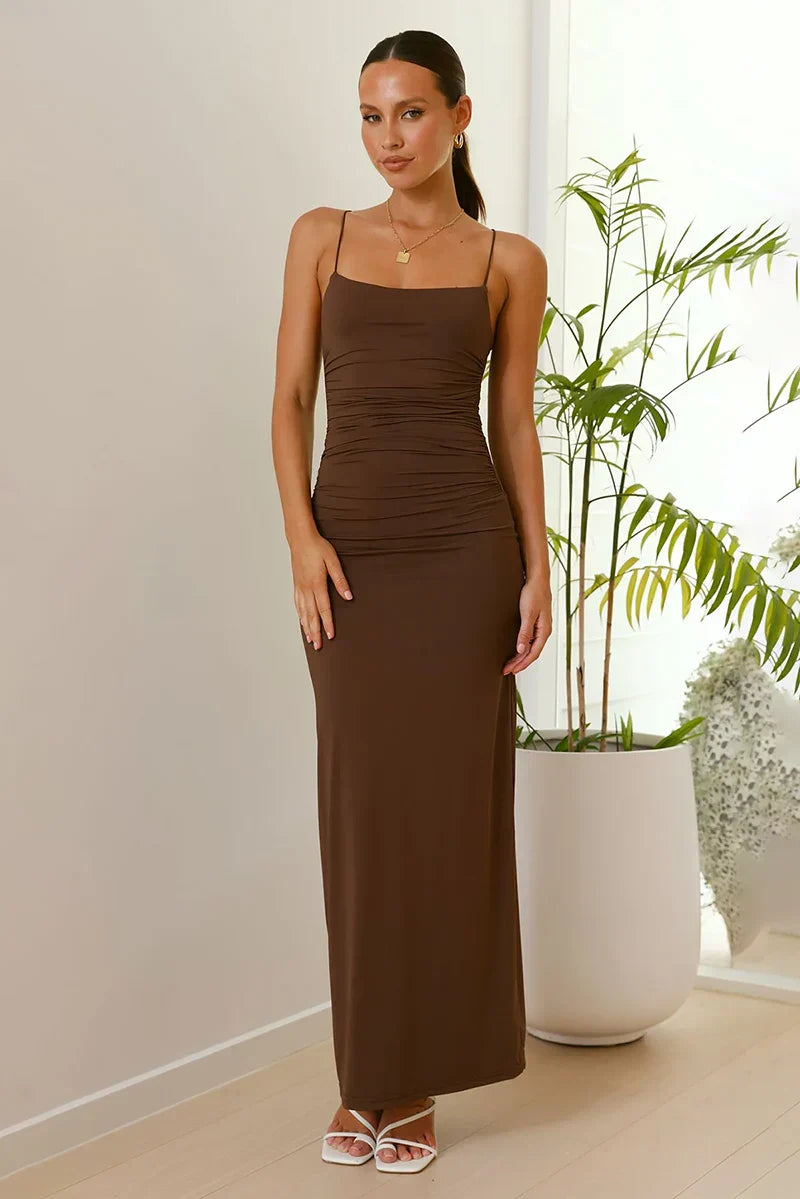 A woman models a form-fitting, solid brown maxi dress with spaghetti straps, a straight neckline, and ruched detailing at the waist.