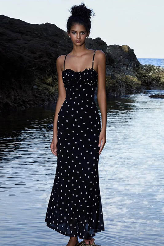 A woman standing by the water wearing a black maxi dress with white polka dots, featuring spaghetti straps and a ruched bodice.