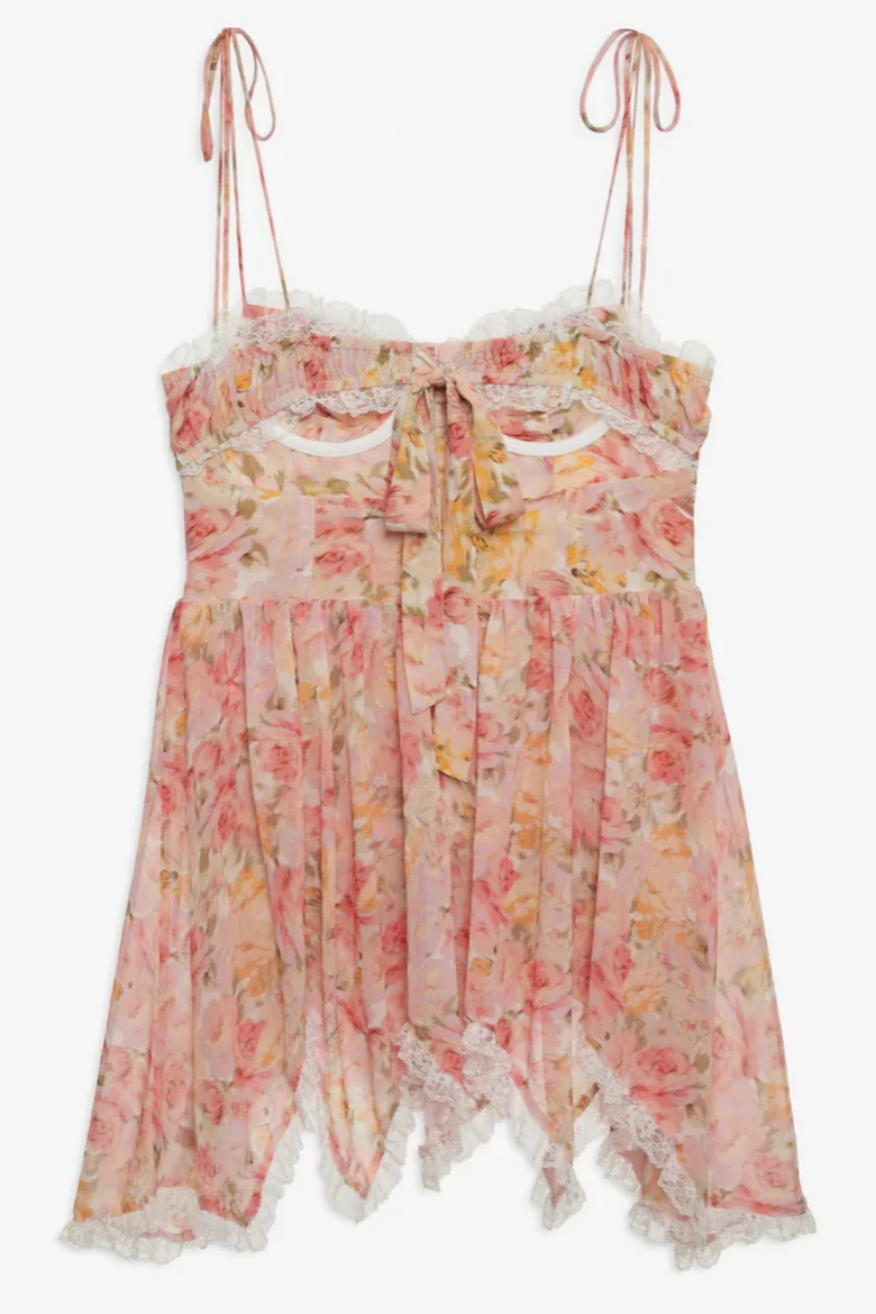 Elea - A woman models a romantic floral mini dress featuring a pale pink and yellow rose print. The dress has a structured corset bodice with a sweetheart neckline, delicate tie-shoulder straps, a rosette appliqué at the bust, and a layered, ruffled asymmetrical skirt trimmed with white lace.