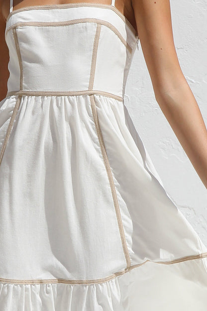White - A woman wearing a white midi dress with thin straps and tan contrast piping on the bodice and tiered skirt. She is smiling and standing against a white textured wall, holding a woven clutch.