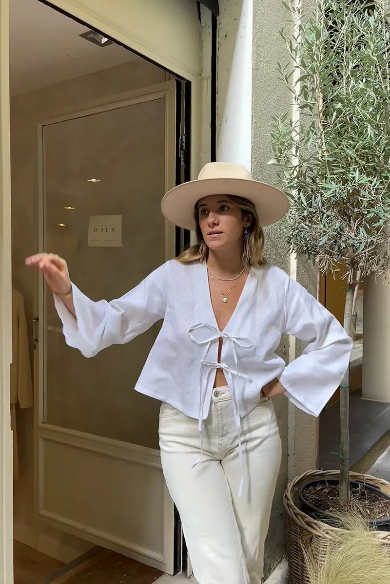 White - Women's white long-sleeve holiday blouse featuring a tie-front closure, V-neckline, and a relaxed, breezy fit, styled with neutral tailored shorts.