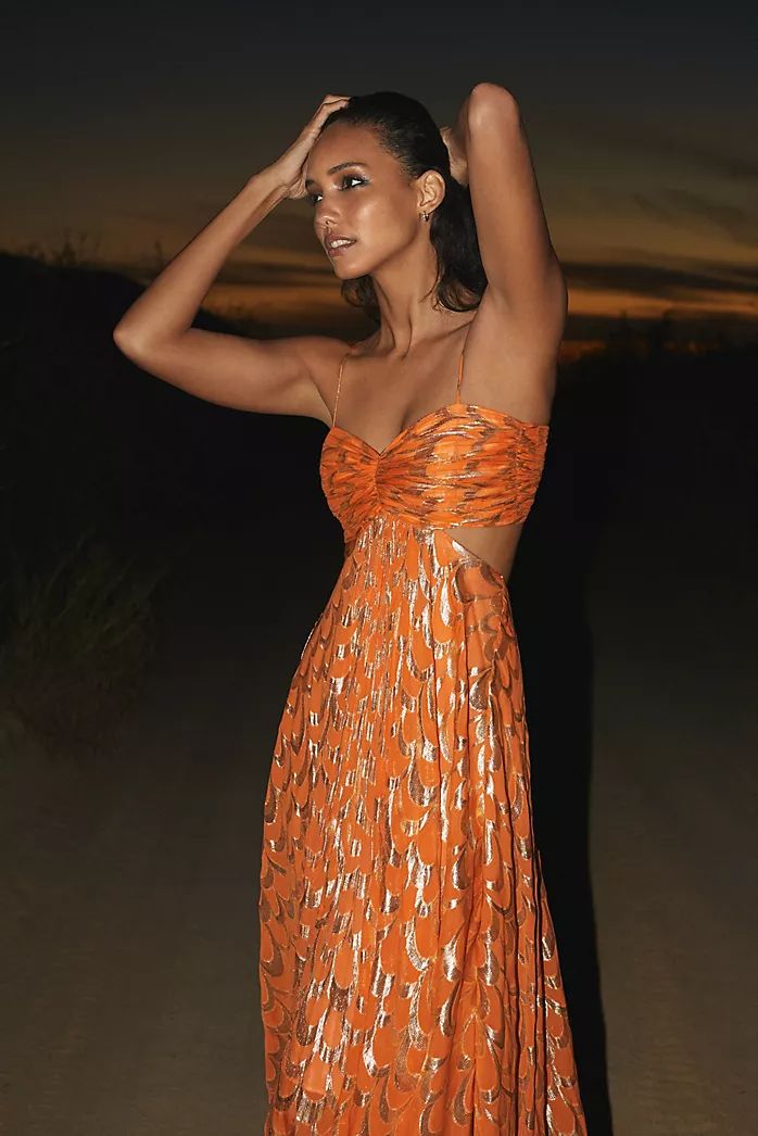 A woman modelling a vibrant orange maxi dress featuring a metallic gold scallop pattern, a ruched sweetheart bodice, side cut-outs, and delicate spaghetti straps.