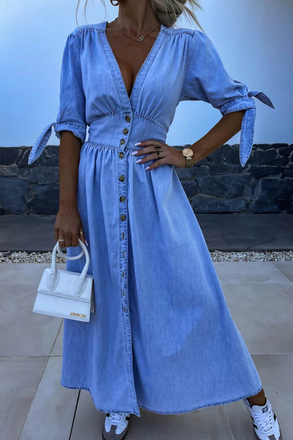 Blue - Woman wearing a light blue denim-look midi dress with a V-neck, button-down front, and tie-detail short sleeves, styled with trainers and a small white handbag.