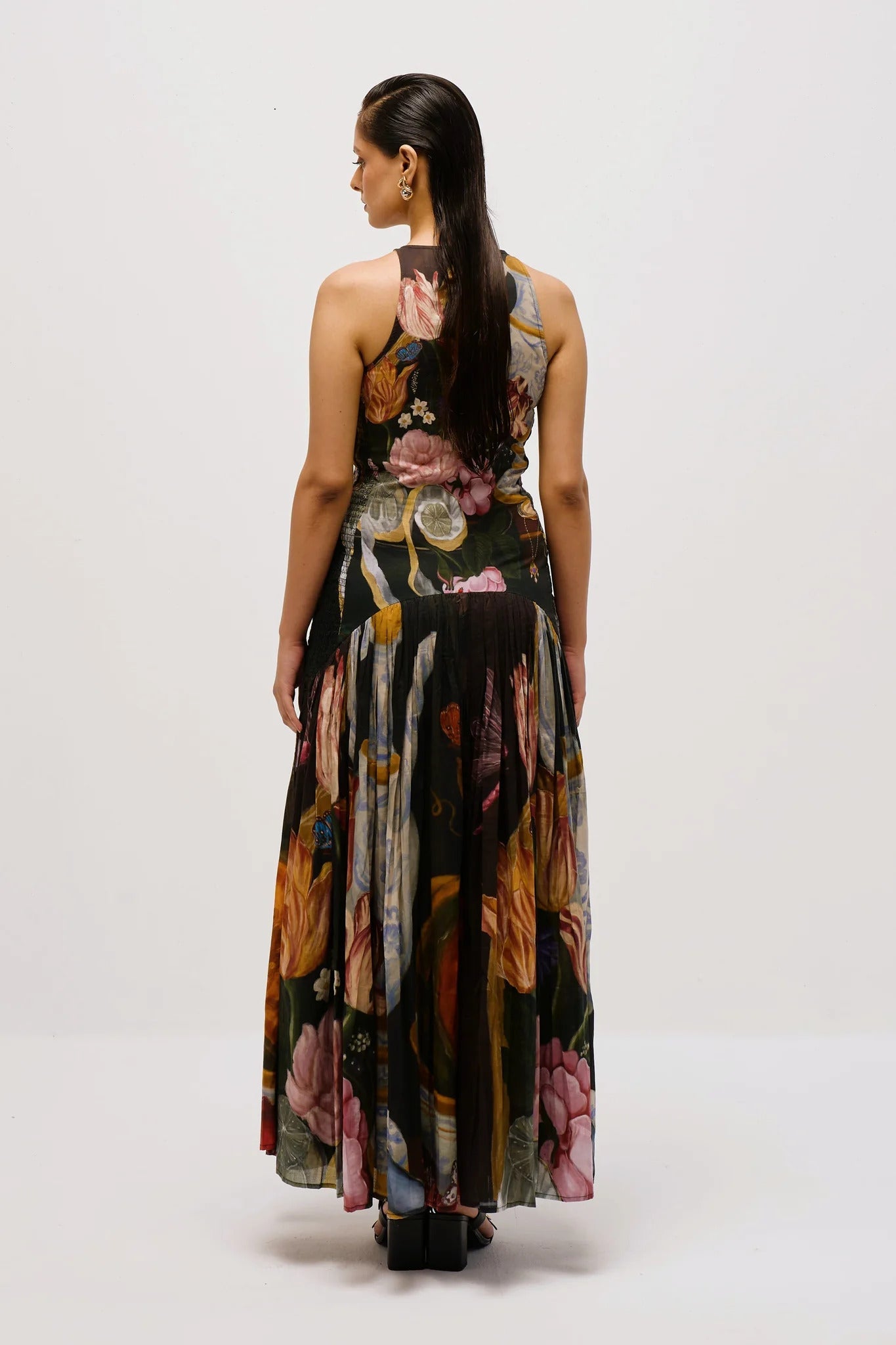 Multi - Full-length view of a woman wearing a sleeveless maxi dress with a high halter neckline and a pleated skirt, featuring a detailed still life floral and vase print on a dark background.