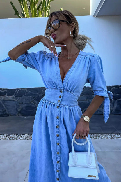 Blue - Woman wearing a light blue denim-look midi dress with a V-neck, button-down front, and tie-detail short sleeves, styled with trainers and a small white handbag.
