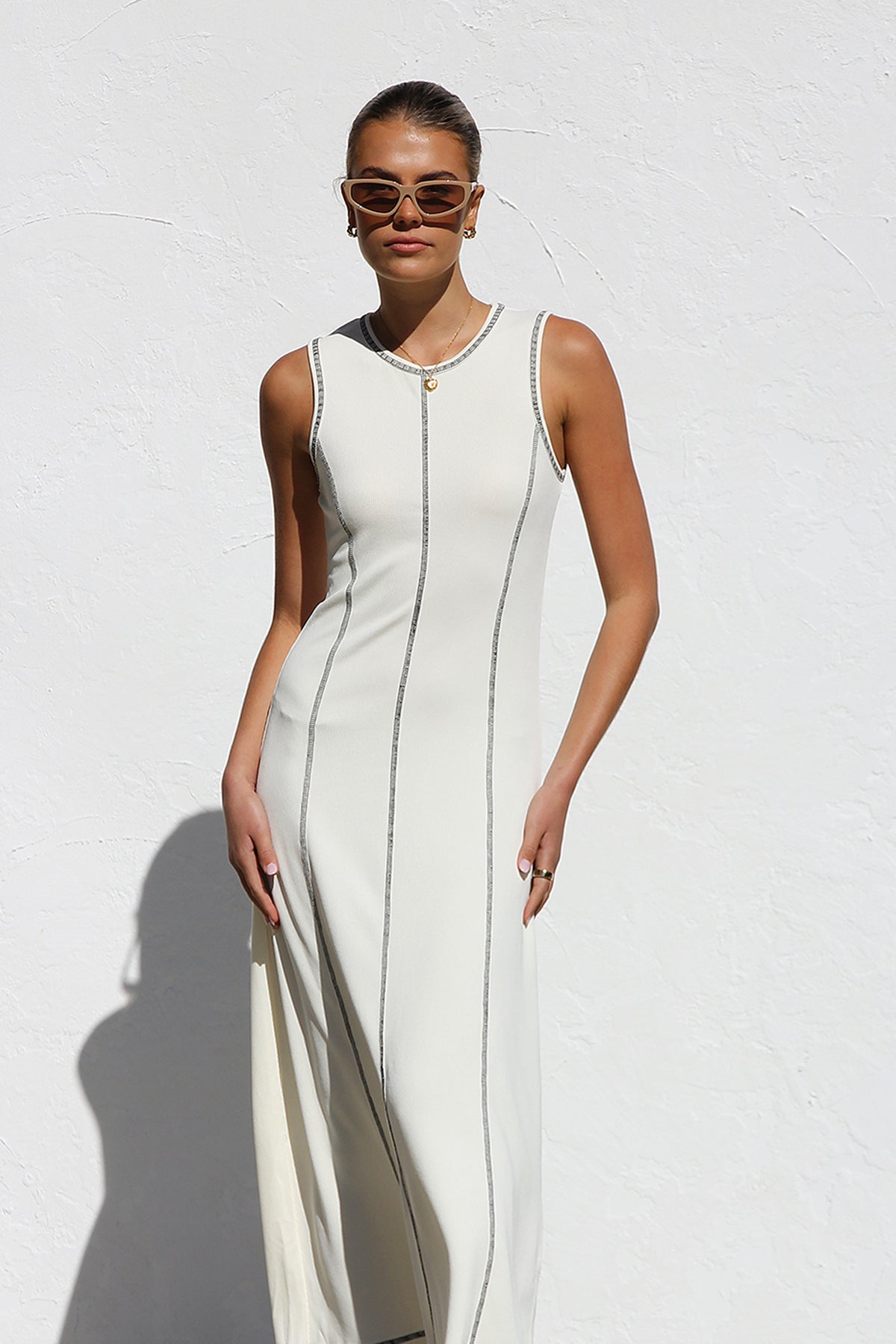 White - A woman stands against a white wall wearing a sleeveless, form-fitting, off-white midi dress. The dress has a high neckline and is detailed with dark vertical contrast stitching along the seams and armholes.