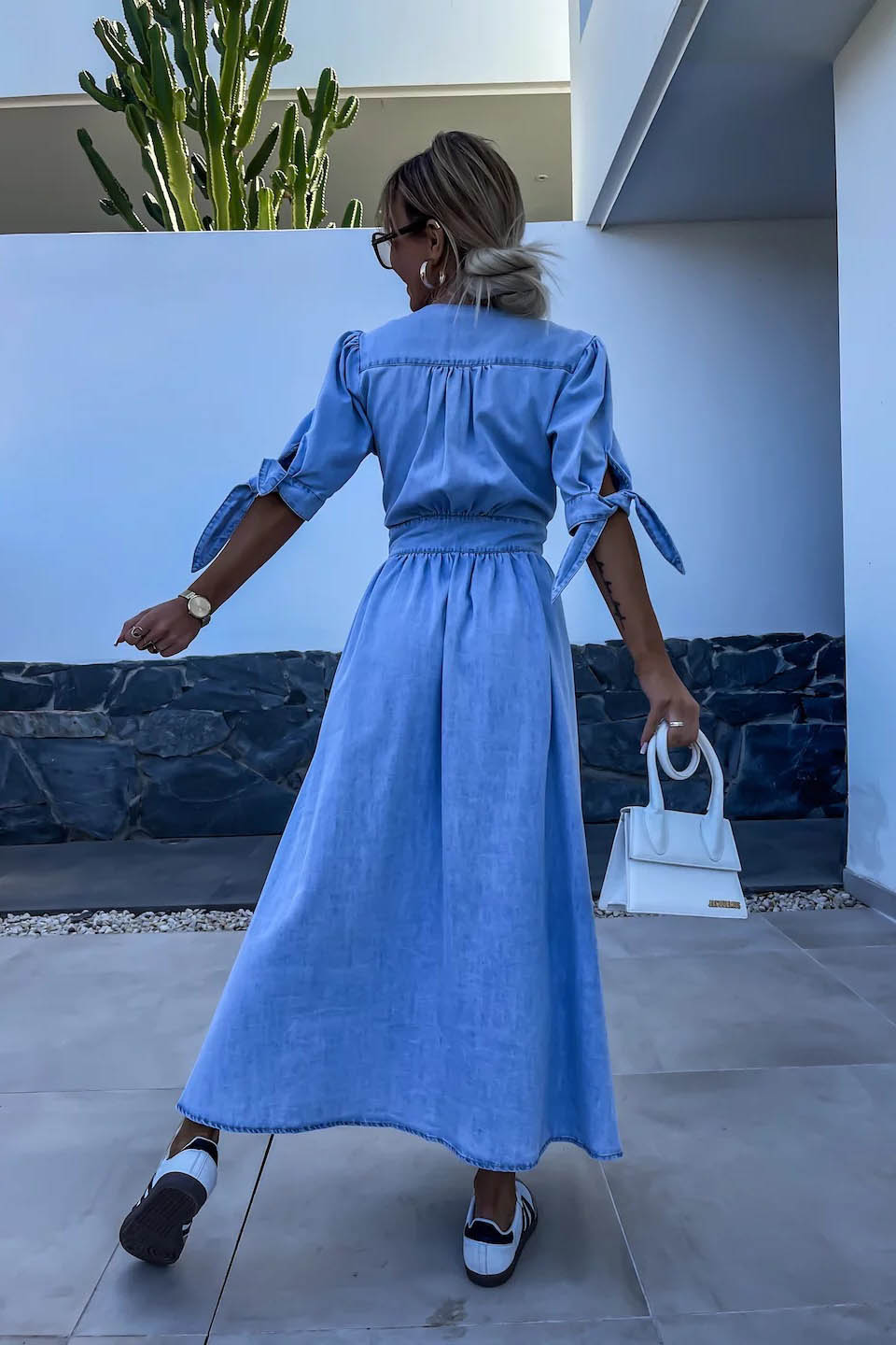 Blue - Woman wearing a light blue denim-look midi dress with a V-neck, button-down front, and tie-detail short sleeves, styled with trainers and a small white handbag.