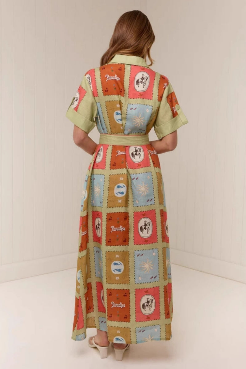 Multicolored - A woman wearing a long, short-sleeved shirt dress featuring a colourful patchwork tile print with whimsical motifs. The dress has a button-down front, a collar, a matching waist belt, and side pockets.