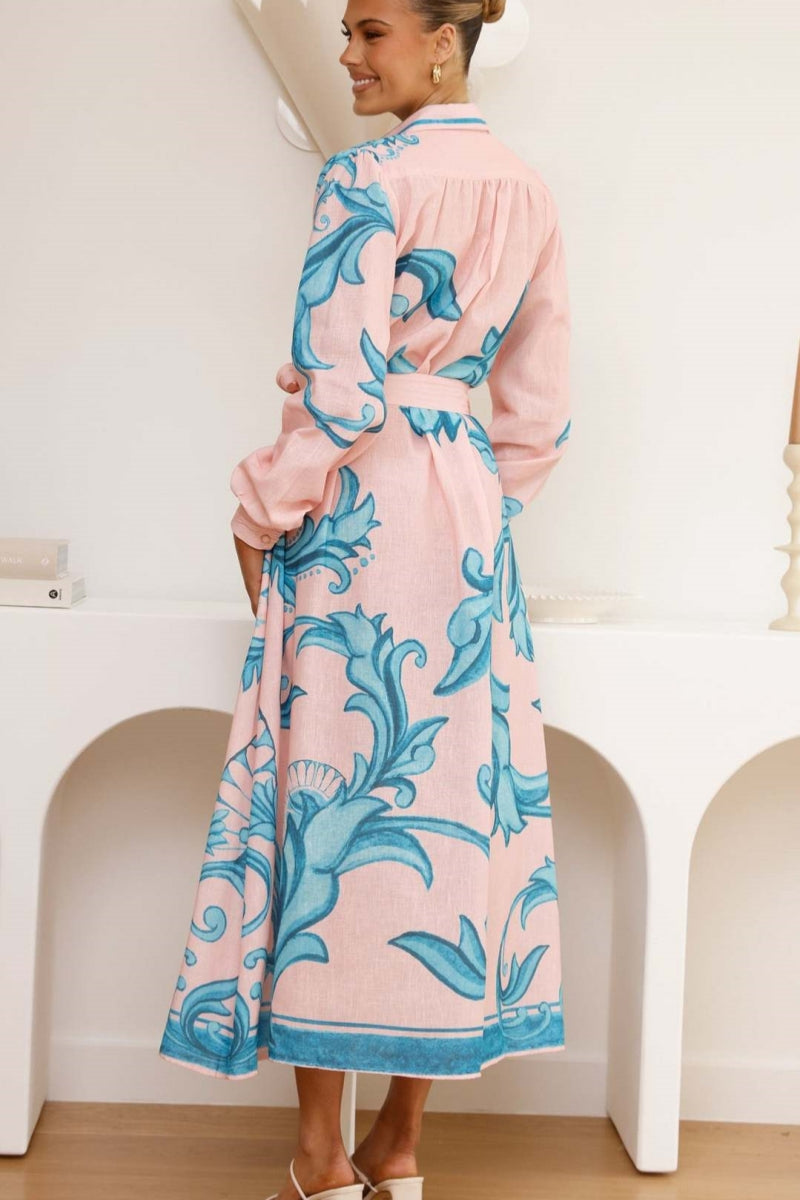 Green - Full-length view of a woman wearing a pink maxi shirt dress featuring a bold blue baroque print, long blouson sleeves, a collared neck, and a matching tie-belt at the waist.