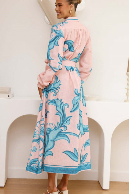 Green - Full-length view of a woman wearing a pink maxi shirt dress featuring a bold blue baroque print, long blouson sleeves, a collared neck, and a matching tie-belt at the waist.