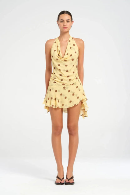 Yellow - A woman wearing a pale yellow halter neck mini dress with large brown polka dots, featuring a cowl neckline, side ruching, and an asymmetrical ruffled hem.